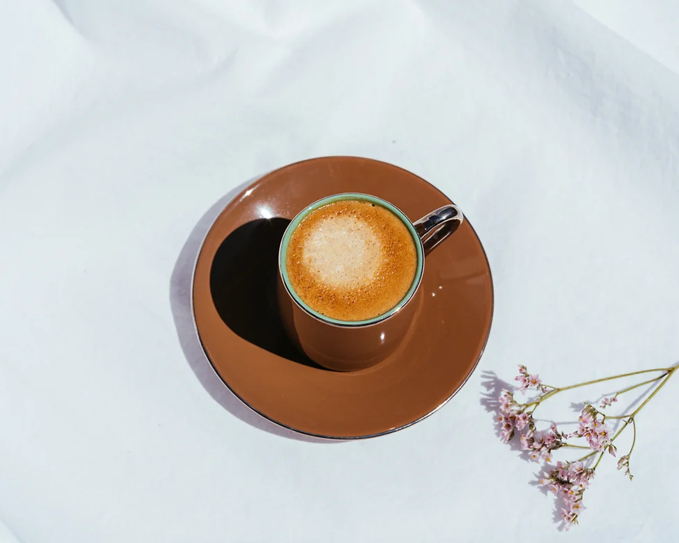 Overhead shot of a frothy latte in a green-rimmed brown cup on a matching saucer, placed on a white cloth with a small pastry nearby