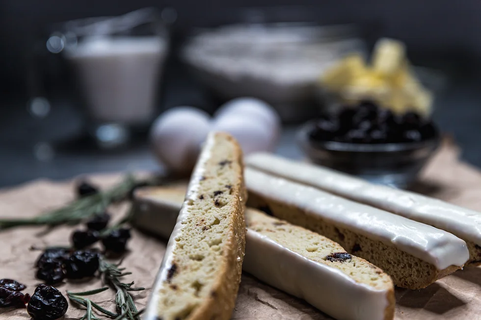 White-glazed biscotti with dried cherries on parchment paper, rosemary sprigs and eggs nearby, soft cream and red tones in a kitchen setting