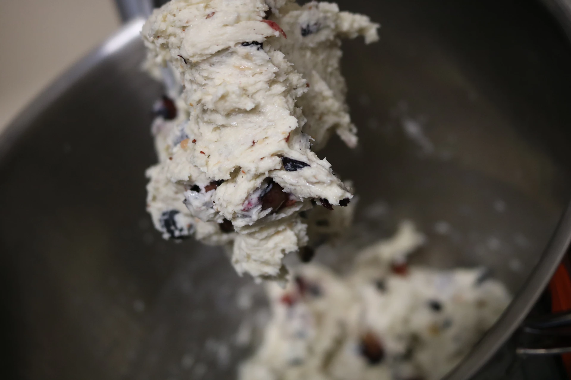 Close-up of creamy dough studded with dark fruit pieces being lifted from a metal mixing bowl, pale and dark tones, kitchen workspace