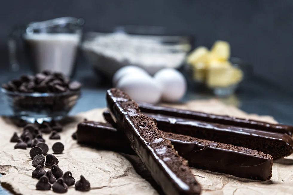 Chocolate-dipped biscotti sticks on parchment paper with chocolate chips scattered beside, eggs and butter blurred in the background, rich brown tones