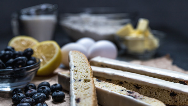 Iced biscotti slices with blueberries and lemon halves, eggs, milk and flour nearby, fresh fruity colors on a light kitchen surface