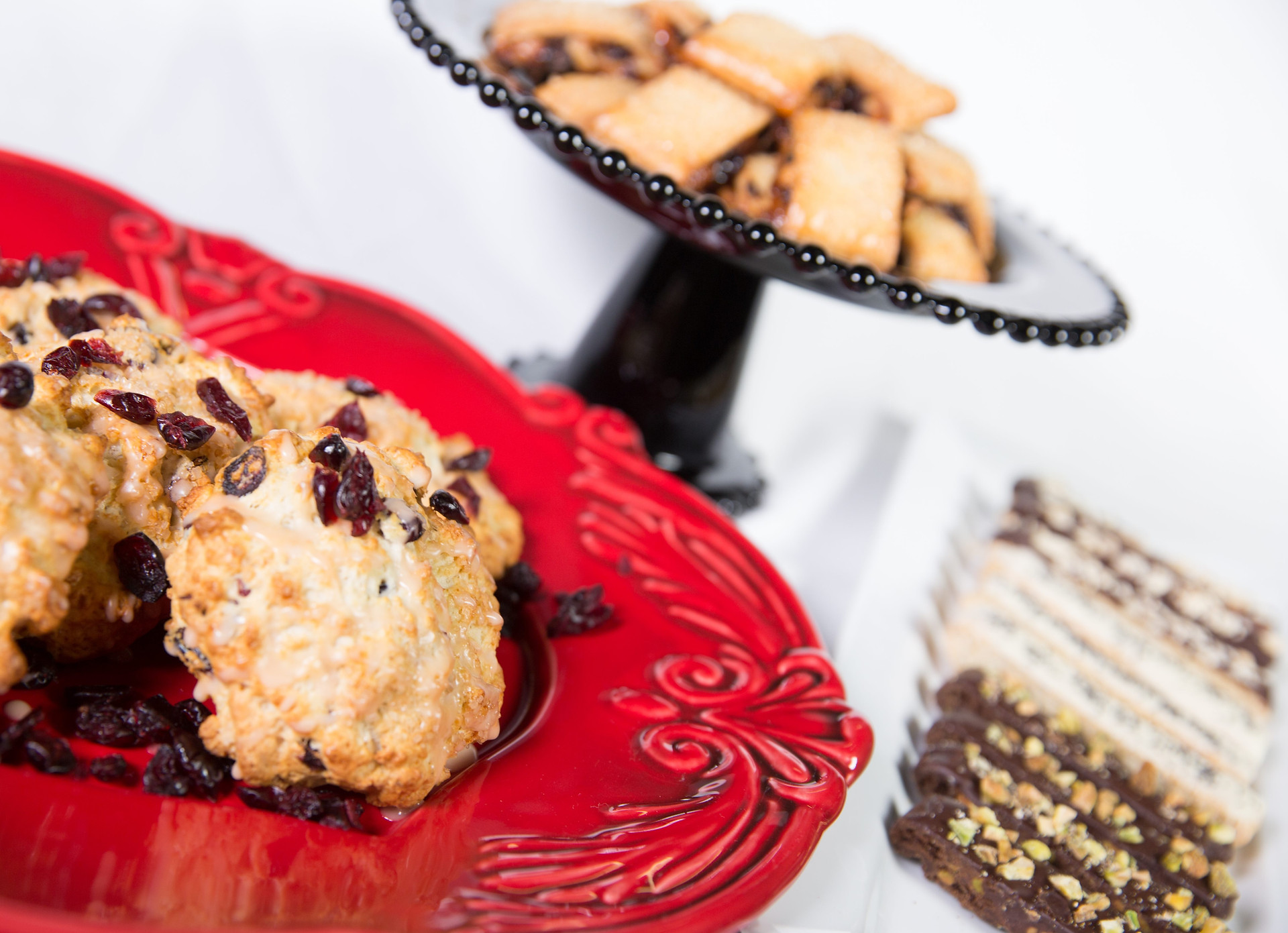 Assorted baked goods including cranberry-topped scones on a red plate and fruit-filled pastries on a black pedestal stand, warm bakery display