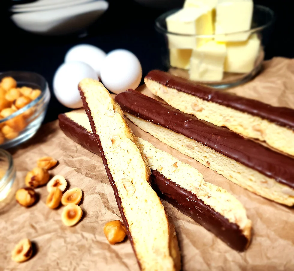 Close-up of chocolate-dipped biscotti sticks on brown parchment paper with scattered hazelnuts, eggs, and a bowl in the background, warm earthy tones